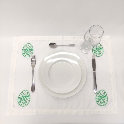 Mesa posta com talheres, pratos brancos e copo sobre individual branco com desenhos verdes.