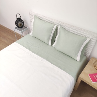 Cama com roupa verde claro e branco, cabeceira trançada branca, mesa de cabeceira com livro e luminária