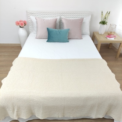 Cama com colcha branca, manta bege, almofadas cinza e azul, cabeceira branca, mesa de madeira com vaso e flores, e vaso no chão com flores