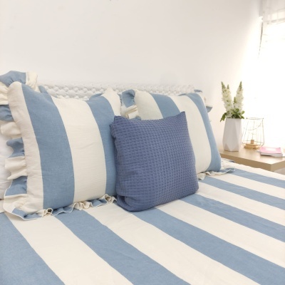 Cama com colcha e almofadas azul claro e branco às riscas com almofada azul escura e mesa de cabeceira com vaso e flores.