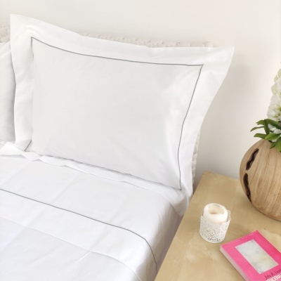 Conjunto de roupa de cama branca com linhas cinzentas simples ao lado de mesa de cabeceira com vaso de madeira, vela e caixa rosa