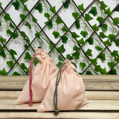 Dois sacos pequenos de tecido cor de rosa com cordões coloridos, sobre banco de madeira com treliça e folhas verdes ao fundo