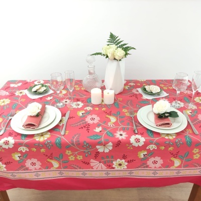 Mesa com toalha vermelha floral, pratos brancos, guardanapos cor de rosa, velas e jarra com flores.