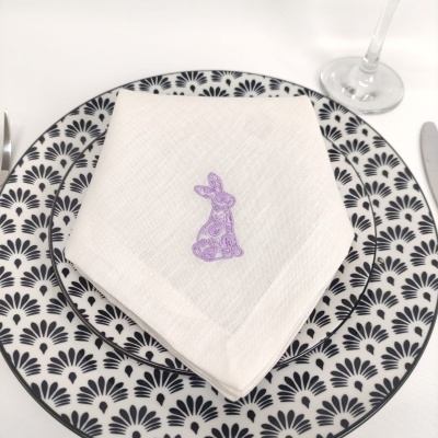 Prato cerâmico branco com padrão preto, guardanapo branco com bordado lilás de coelho, talheres e copo de vidro