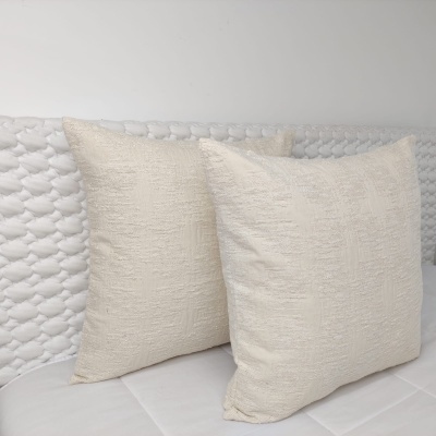 Duas almofadas quadradas bege claro com textura colocadas numa superfície branca acolchoada.