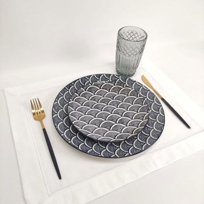 Conjunto de pratos azuis e brancos com padrão de escamas, copo de vidro e talheres dourados e pretos sobre suporte branco