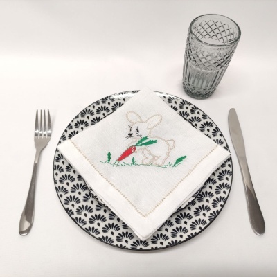 Guardanapo branco com bordado de coelho sobre prato decorado em preto e branco, com talheres e copo de vidro