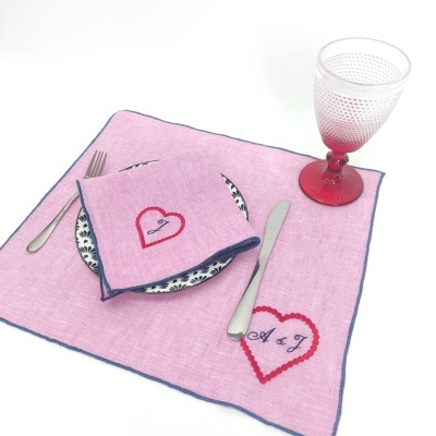 Guardanapo e toalhete rosa com bordados em corações vermelhos e letras, prato azul e branco, copo rosa e talheres