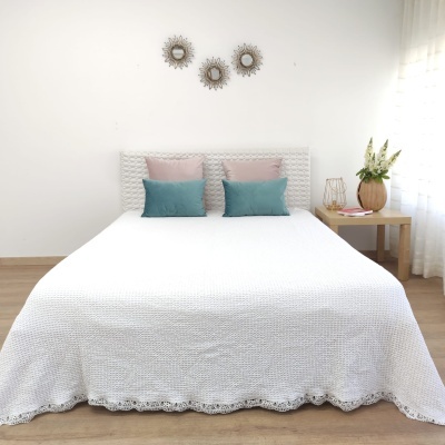 Cama de casal com colcha branca e almofadas azuis e rosas junto a mesa com vaso e luz natural