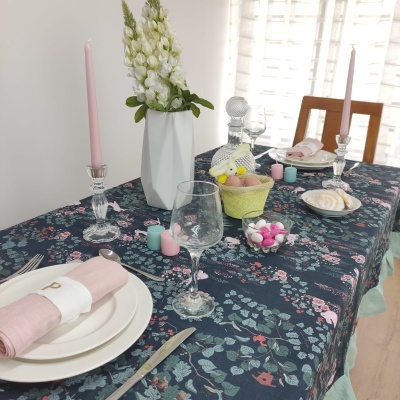 Mesa posta com toalha floral, pratos e copos, velas rosas e vaso com flores brancas