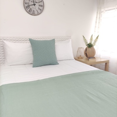 Quarto com cama arrumada com almofadas e cobertor verde-água, relógio na parede e mesa de cabeceira com decoração