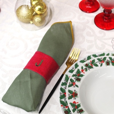 Guardanapo verde com argola vermelha e letra J perto de pratos com padrão de azevinho e copos vermelhos numa mesa decorada