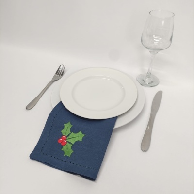 Conjunto de mesa com prato branco, guardanapo azul com bordado de folhas verdes e bagas vermelhas, talheres metálicos e taça de vidro transparente.