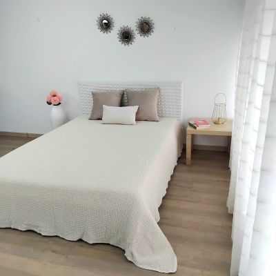 Cama de casal com colcha bege, almofadas castanho e bege, cabeceira branca, mesa com livros e candeeiro, jarro com flores, espelhos decorativos