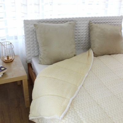 Cobertor bege felpudo sobre cama com cabeceira branca trançada e duas almofadas