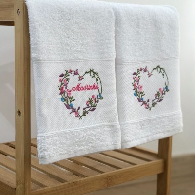 Toalhas brancas com bordado colorido em forma de coração, uma com a palavra Madrinha, penduradas em suporte de madeira.