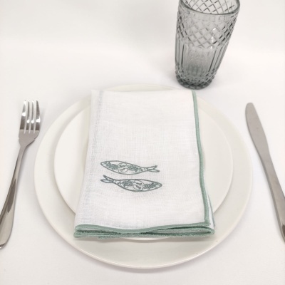 Mesa posta com pratos brancos, guardanapo branco com peixes verdes, talheres metálicos e copo de vidro cinzento.