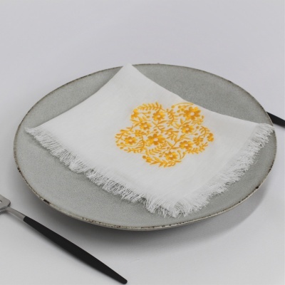 Guardanapo branco com estampa floral amarela sobre prato cinza claro com talheres pretos.