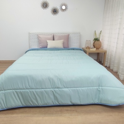 Colcha azul claro em cama branca com cabeceira entrelaçada e almofadas cor-de-rosa e branca, mesa de madeira e vaso decorativo