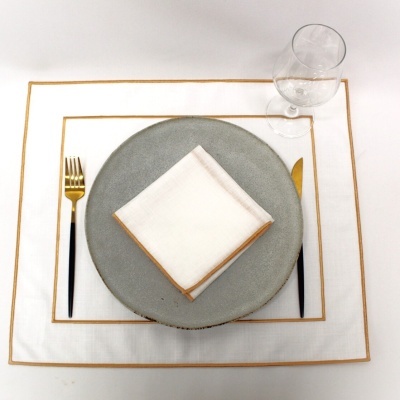 Conjunto de mesa com prato cinza, guardanapo branco com borda dourada, talheres dourados com cabo preto e copo transparente