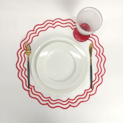 Conjunto de jantar com pratos brancos, talheres dourados com cabos pretos, copo de vidro com base vermelha e toalhete branco com ondas vermelhas.