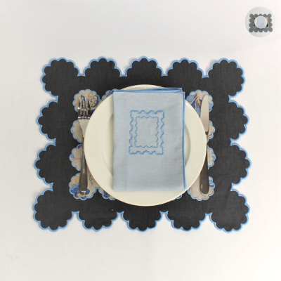 Individual preto com bordas florais, prato branco com guardanapo azul bordado e talheres.