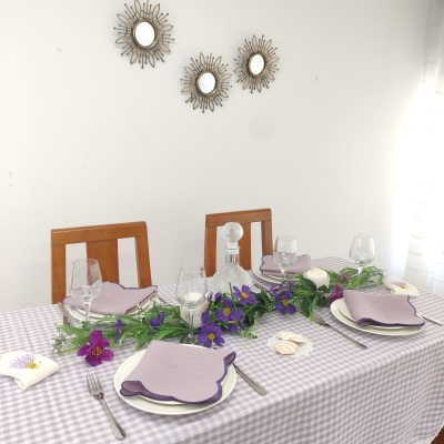Mesa de jantar decorada com toalha xadrez lilás, guardanapos lilás, arranjo floral e vidro de cristal.