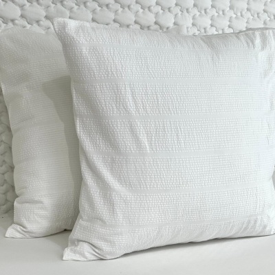 Dois almofadas brancas texturizadas em relevo, sobre superfície e parede brancas com padrão.