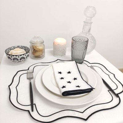 Conjunto de jantar com pratos brancos, guardanapo com estrelas pretas, talheres pretos, copo cinzento, suporte de vidro, vela decorativa e garrafa de vidro.