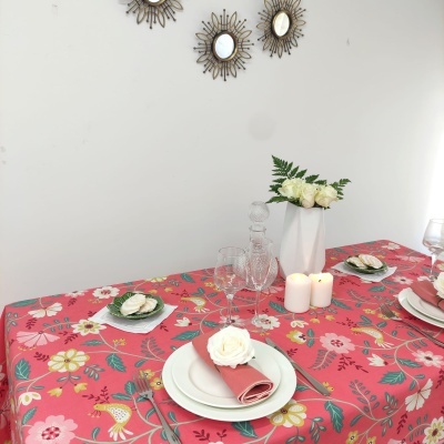 Mesa posta com toalha rosa floral, pratos brancos, guardanapos rosa, velas e flores em jarro branco. Três espelhos dourados na parede branca.