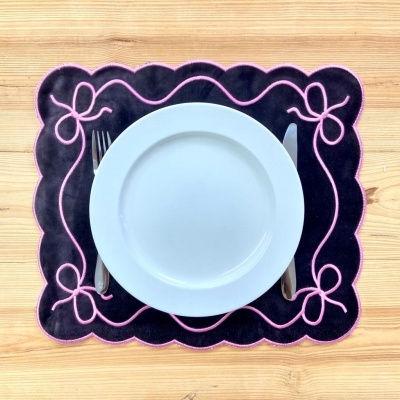 Lugar americano preto com bordado rosa, prato branco, talheres à esquerda e direita, mesa de madeira clara