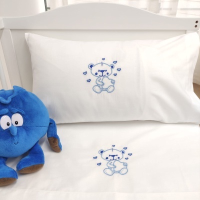 Capa de almofada e lençol bordados com urso azul numa cama branca com pelúcia azul