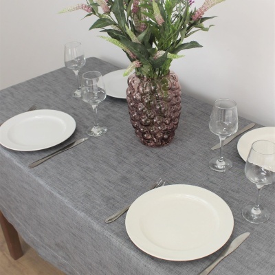 Toalha de mesa cinza com pratos brancos, talheres, copos de vinho e vaso rosa com flores.