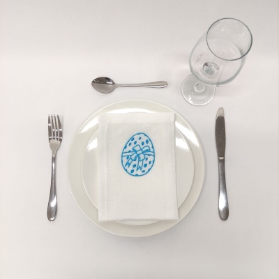 Conjunto de jantar com base branca, guardanapo com desenho azul de ovo, talheres metálicos e copo de vidro