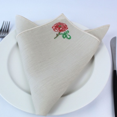 Guardanapo bege com bordado de flor vermelha sobre prato branco com talheres.