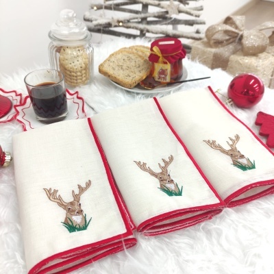 Três panos de cozinha brancos com bordado de veado castanho e detalhes vermelhos, decorados com itens de Natal e pão