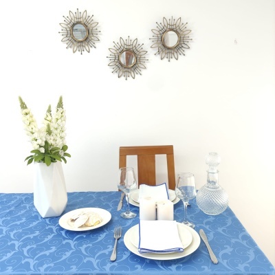 Mesa posta com toalha azul, pratos, copos, vela e vaso branco com flores, cadeira de madeira e três espelhos decorativos na parede