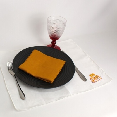 Conjunto de louça com prato preto, guardanapo mostarda, talheres prateados, copo vermelho texturizado e toalha branca com bordado.