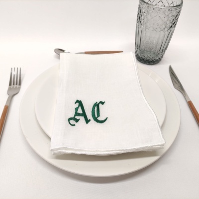 Guardanapo branco com bordado verde AC sobre prato branco com talheres e copo de vidro.