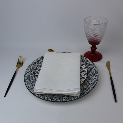 Conjunto de pratos decorativos branco com padrão preto, talheres com cabos dourados e pretos, guardanapo branco e copo vermelho translúcido texturizado