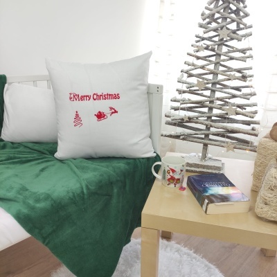 Almofada branca 'Merry Christmas' num sofá com manta verde e mesa com caneca, livro e árvore decorativa