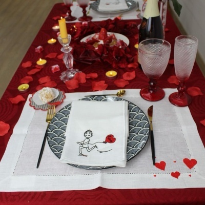 Lugar de mesa decorado com guardanapo branco bordado, prato cinzento, talheres dourados, copos de vidro e decoração de pétalas vermelhas e velas.