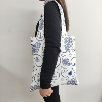 Mala tote branca com padrão azul em espiral e flores, segurada ao lado por pessoa com camisola preta