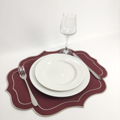 Conjunto de jantar com pratos brancos, talheres metálicos, copo de vinho e sousplat bordô com bordas recortadas.