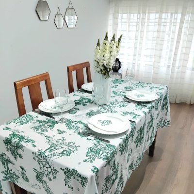 Mesa de jantar com toalha branca e padrão verde, pratos e copos, vaso com flores, cadeiras de madeira e três espelhos na parede
