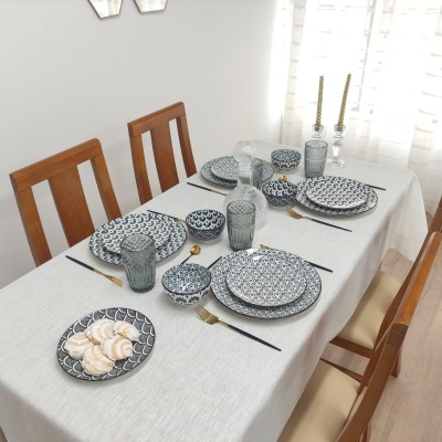 Mesa posta com conjunto de louça preta e branca, copos texturizados, talheres pretos e dourados, velas douradas em castiçais, toalha branca e cadeiras de madeira