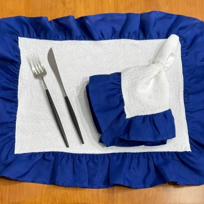 Conjunto de mesa com individual branco e azul, guardanapo branco com detalhe azul, garfo e faca com cabos pretos