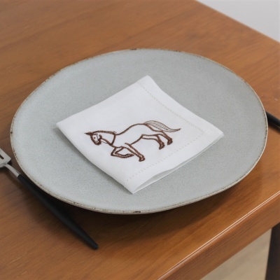 Guardanapo branco bordado com cavalo sobre prato cinza e talheres pretos numa mesa de madeira.