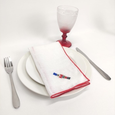 Conjunto de loiça com pratos brancos, guardanapo branco com borda vermelha, talheres metálicos e copo com haste vermelha