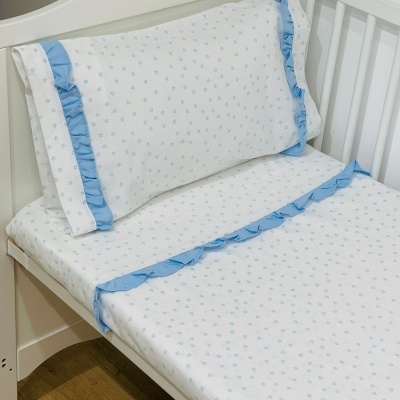 Lençol e fronha brancos com flores azuis e folhos azuis numa cama de grades branca